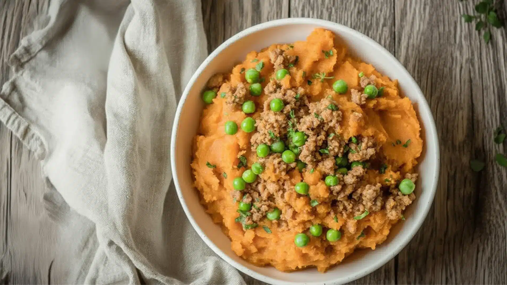 A bowl of savory sweet potato mash topped with seasoned ground meat and green peas, served on a rustic wooden table