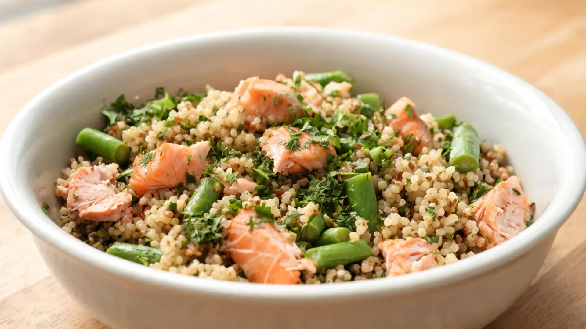 A white bowl filled with salmon pieces, green beans, and fresh herbs on a wooden surface